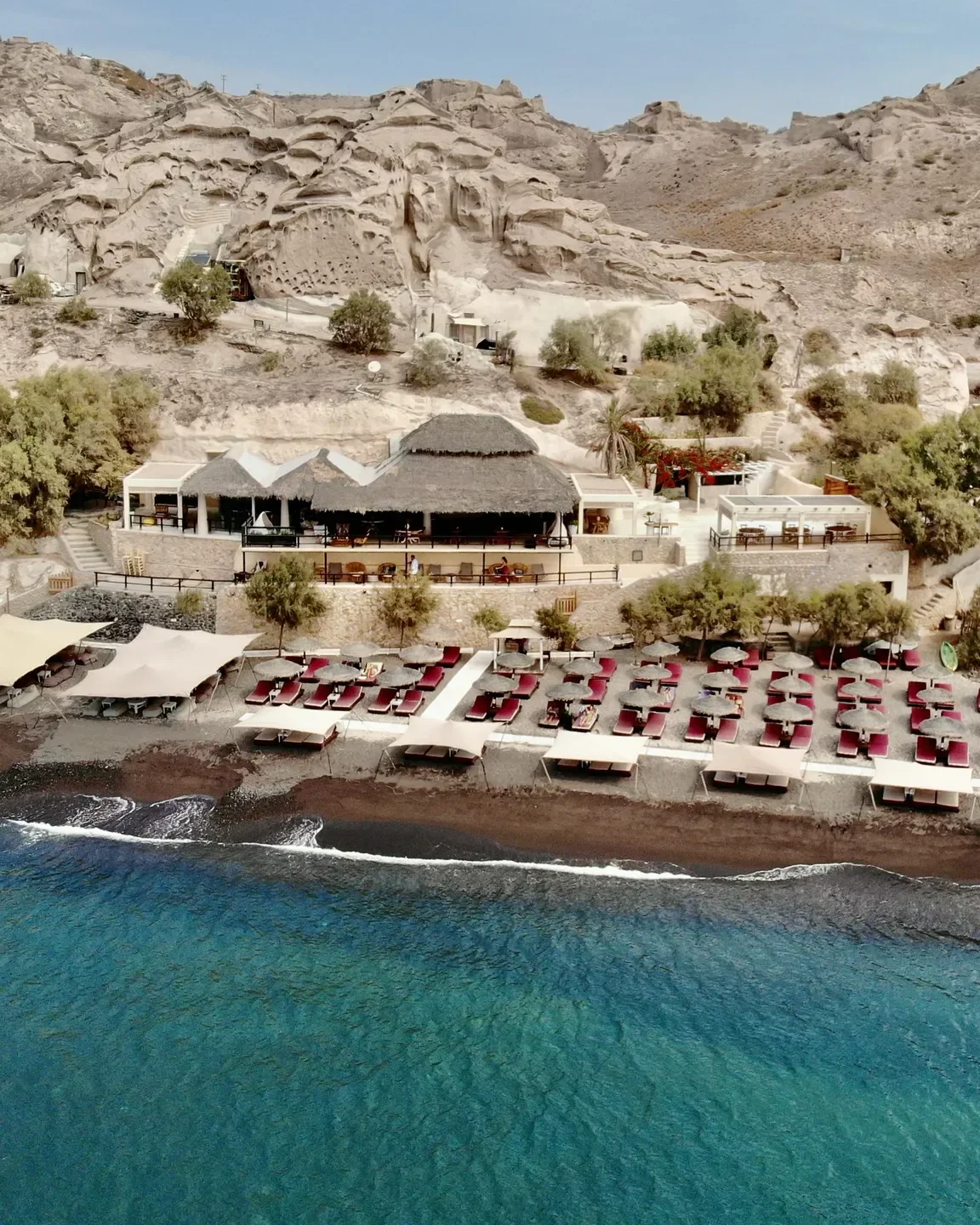 Beachfront view of Theros Wave Bar in Santorini with sunbeds and turquoise waters.